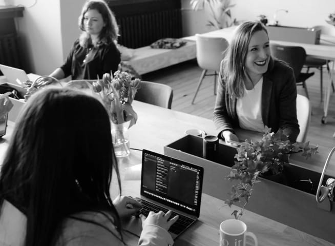 Image of a work team: two of the three women are concentrating using their laptops, while one of them smiles and holds a phone in her hand.